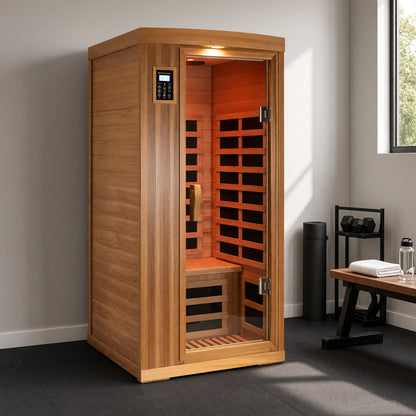 One-person red cedar infrared sauna in a home gym on rubber flooring beside a dumbbell rack and a bench with a towel